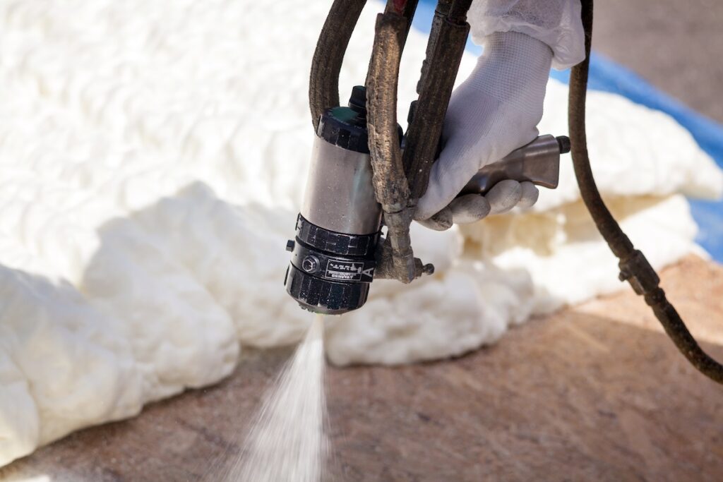 A gloved hand operates a spray foam gun, applying white insulation foam onto a surface, with cured foam visible in the background. diy spray foam insulation