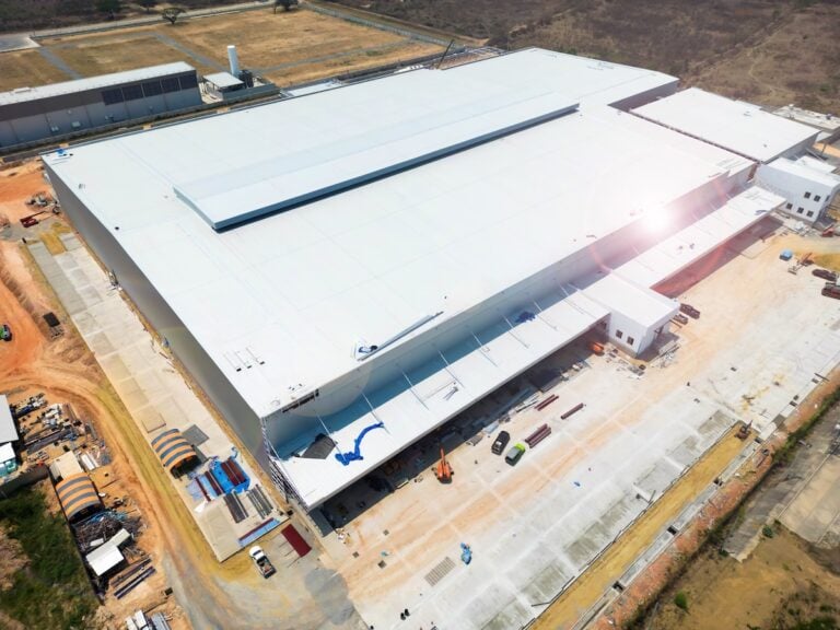 Aerial view of a large, white-roofed industrial warehouse under construction, surrounded by construction equipment, vehicles, and materials, with some areas dirt and others paved.