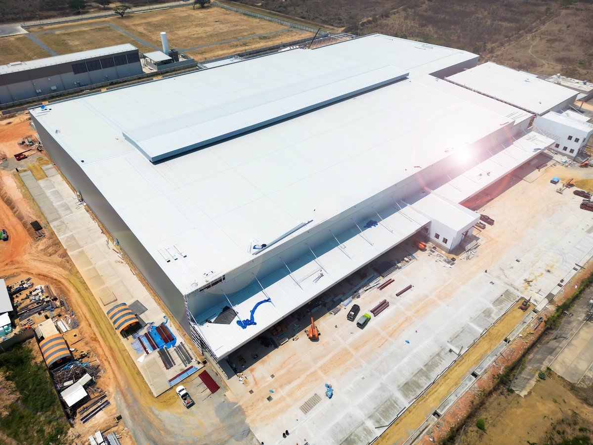Aerial view of a large, white-roofed industrial warehouse under construction, surrounded by construction equipment, vehicles, and materials, with some areas dirt and others paved.