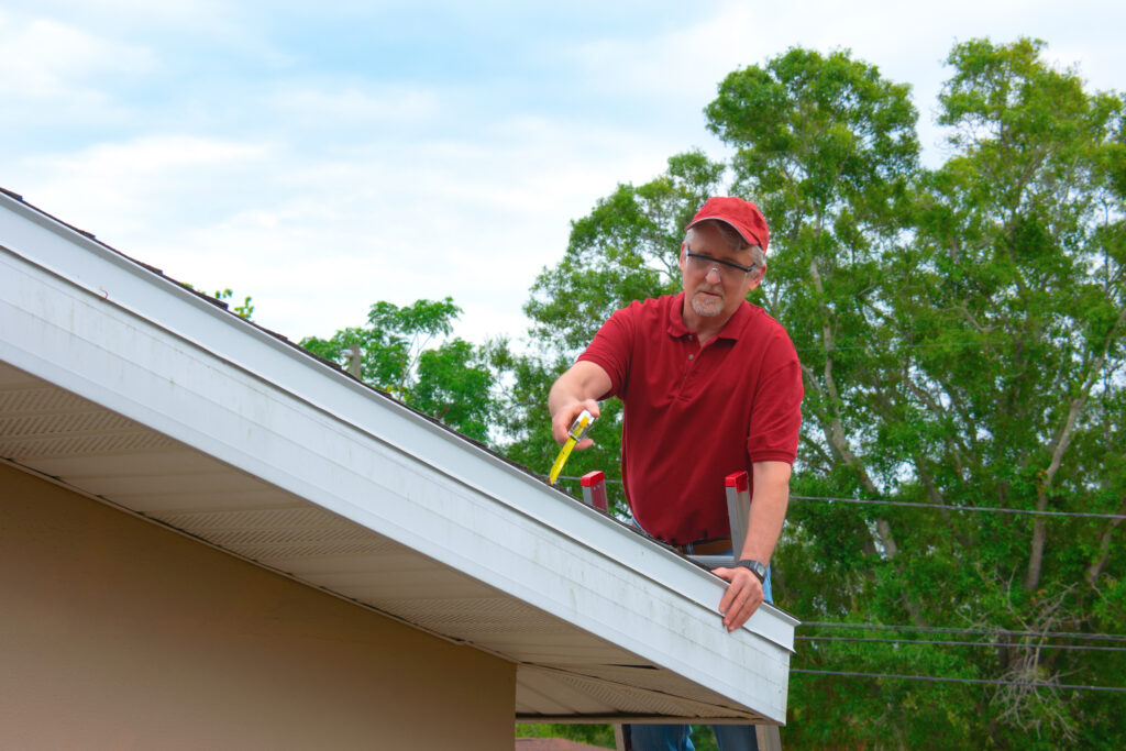 A man in a red shirt and cap cleans leaves from a house gutter using a tool while standing on a ladder. Green trees and a partly cloudy sky are in the background. soffit repair