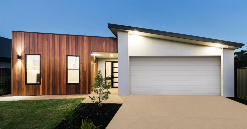 Modern single-story flat roof house with a wooden and white exterior, large windows, a double garage door, a small front yard with green grass, and outdoor lights illuminating the entrance at dusk.