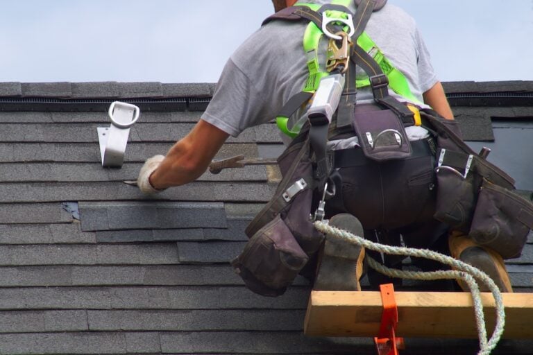 A construction worker wearing safety gear and a harness performs shingle roof repair, installing asphalt shingles on a roof while using tools and standing on a wooden platform secured by ropes.