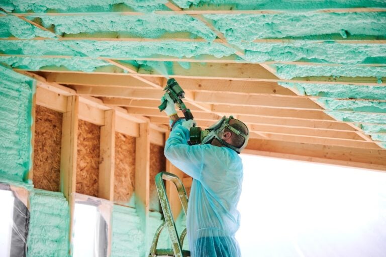 A worker in protective gear stands on a ladder, spraying green foam insulation onto the wooden ceiling beams of a building under construction. Bright light shines through windows in the background.