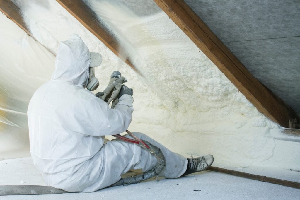 A worker in protective gear sprays foam insulation onto the interior roof of an attic, sitting on the floor with equipment and hoses. diy spray foam insulation