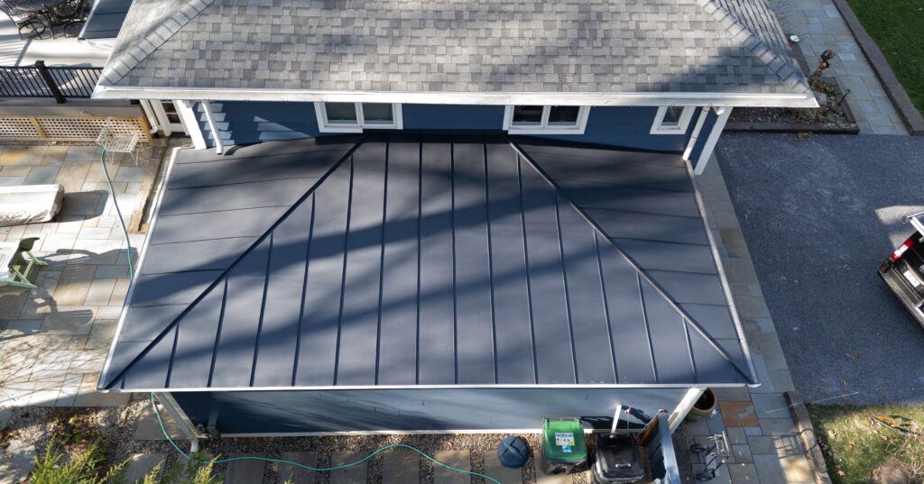 Aerial view of a house with a gray standing seam metal roof attached to a blue building. Concrete patio, driveway, and parked vehicles surround the structure—showcasing why many wonder: are metal roofs better for durability and style? Trees cast shadows overhead.
